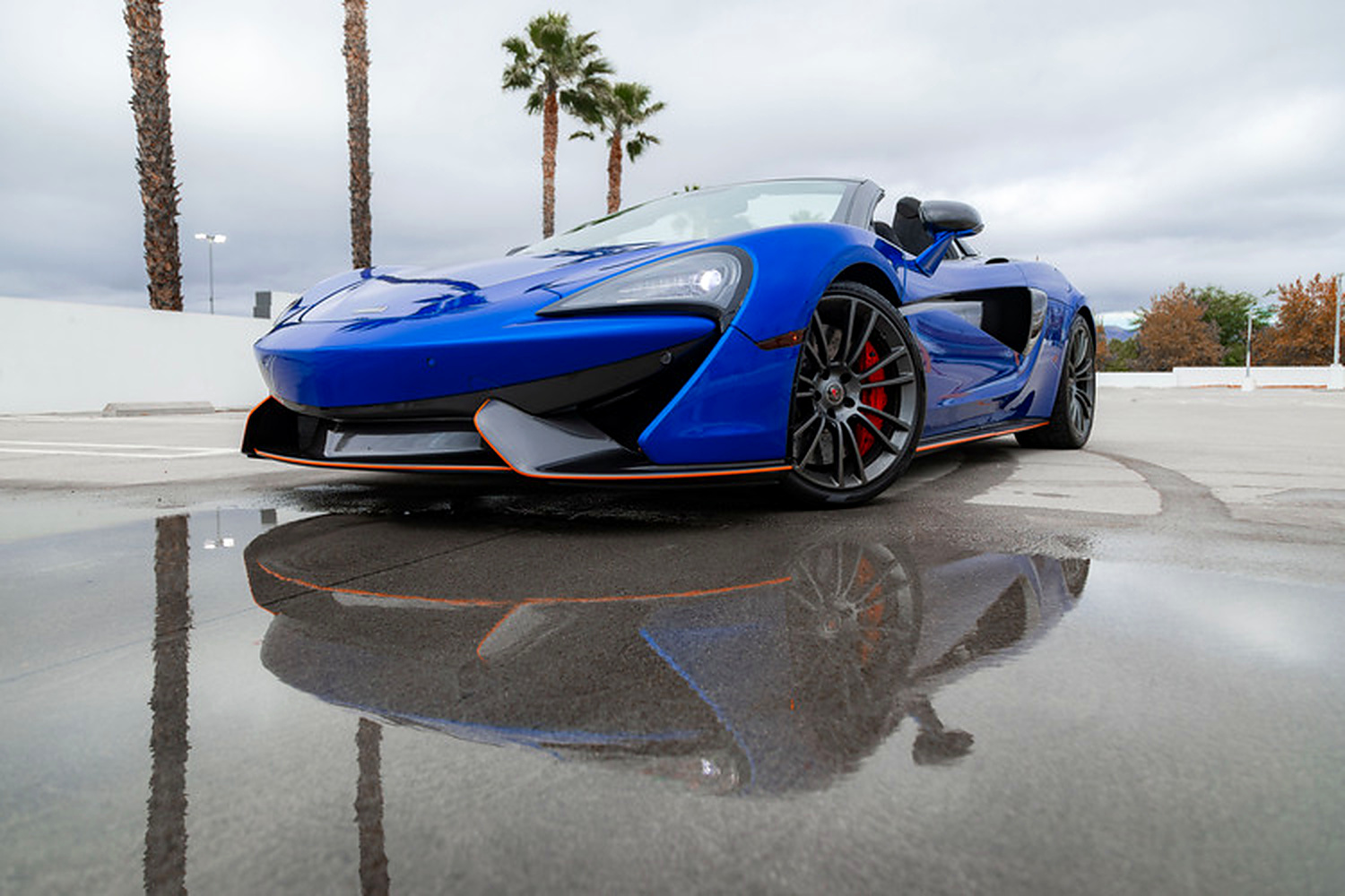 Head-on view of the blue 570S Spider reflected in wet pavement, orange splitter line and the roof down