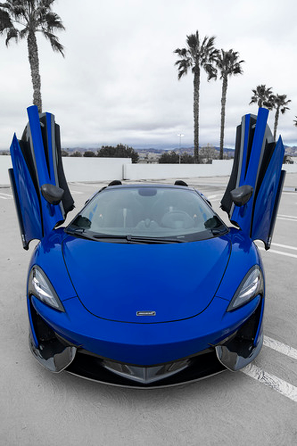 Blue McLaren 570S Spider with both dihedral doors raised and the roof folded, palm trees behind