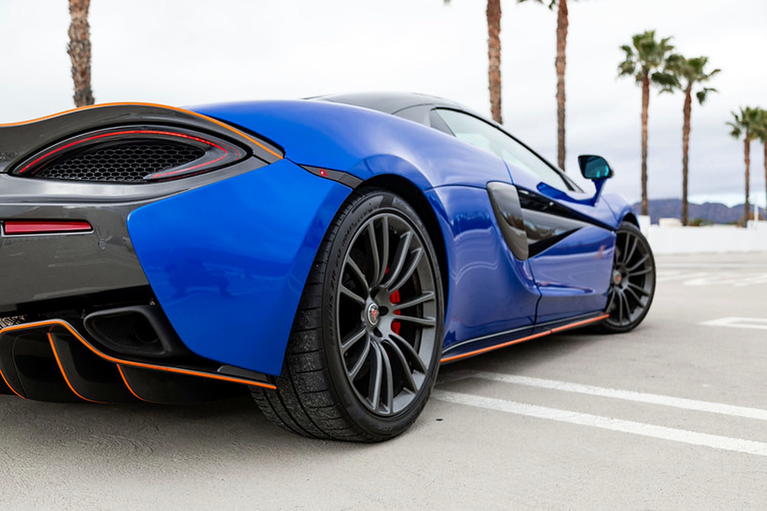 Rear three-quarter of the Vega Blue 570S Spider, dark multi-spoke wheels, red calipers and orange diffuser line