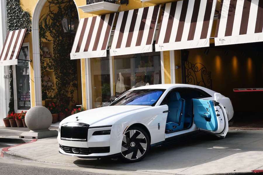 White Rolls Royce Spectre with its coach door open on a turquoise cabin outside a striped-awning storefront on Rodeo Drive
