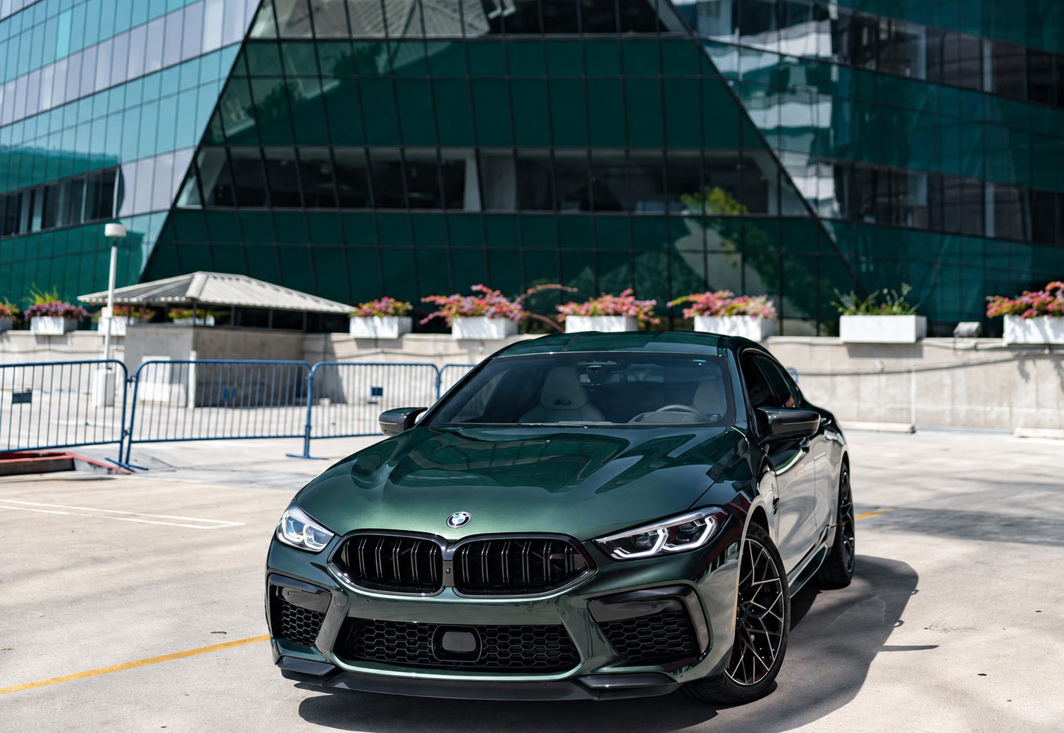Low front view of the dark green BMW M8 Competition Gran Coupe with a glass building behind
