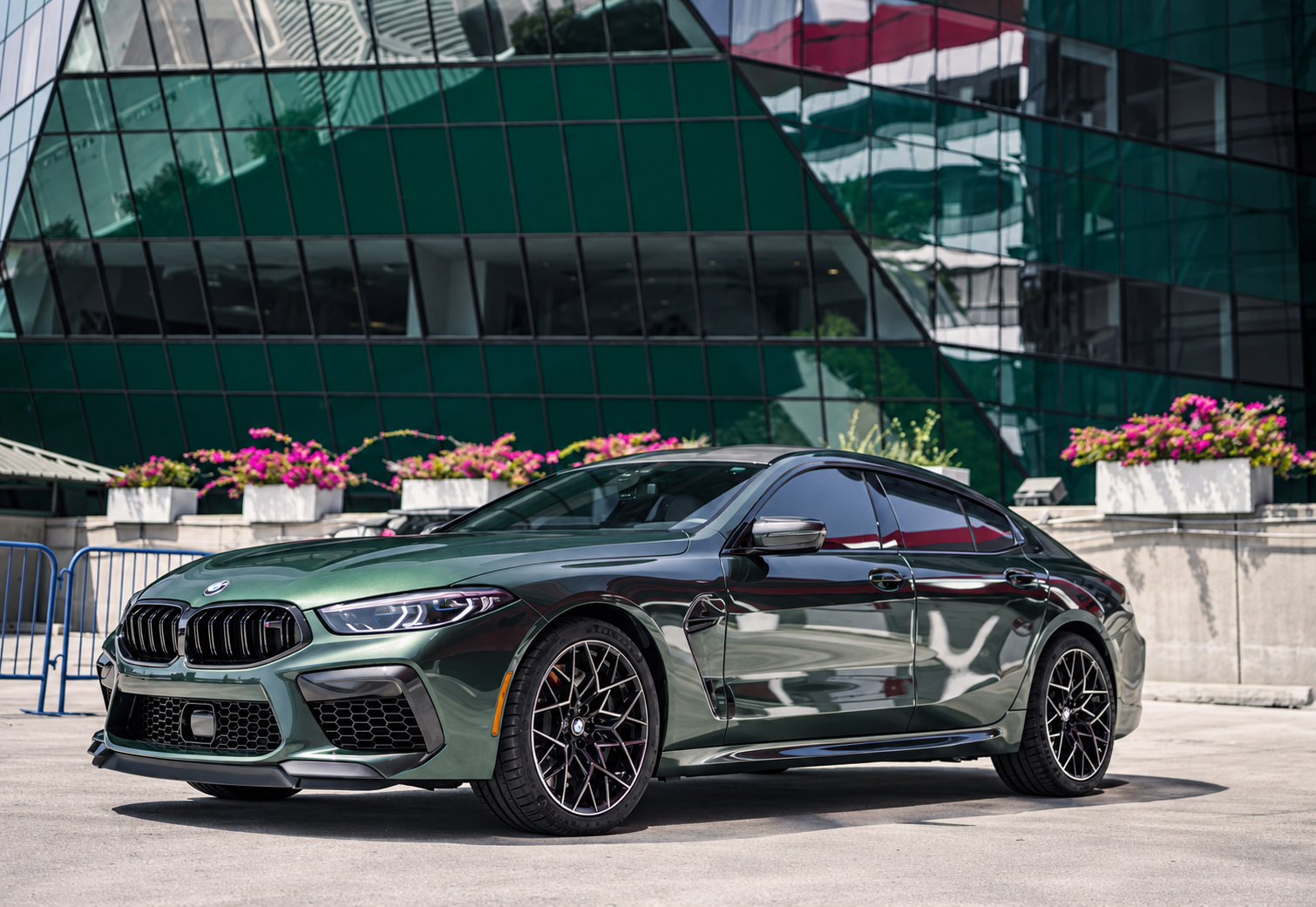 Dark green BMW M8 Competition Gran Coupe front three-quarter outside a green glass office building