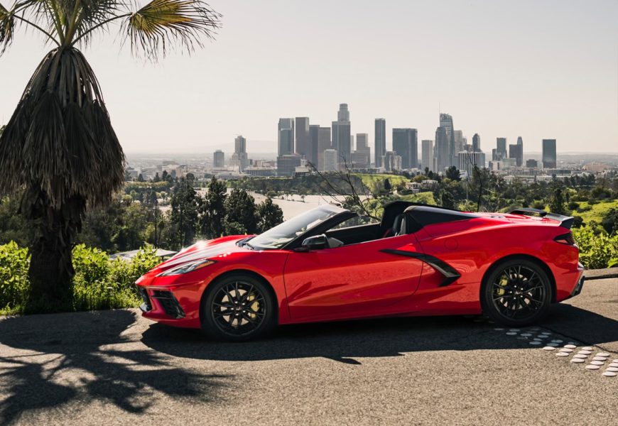 Side profile of the red C8 Stingray, roof off, black wheels and rear spoiler, with the Los Angeles skyline behind