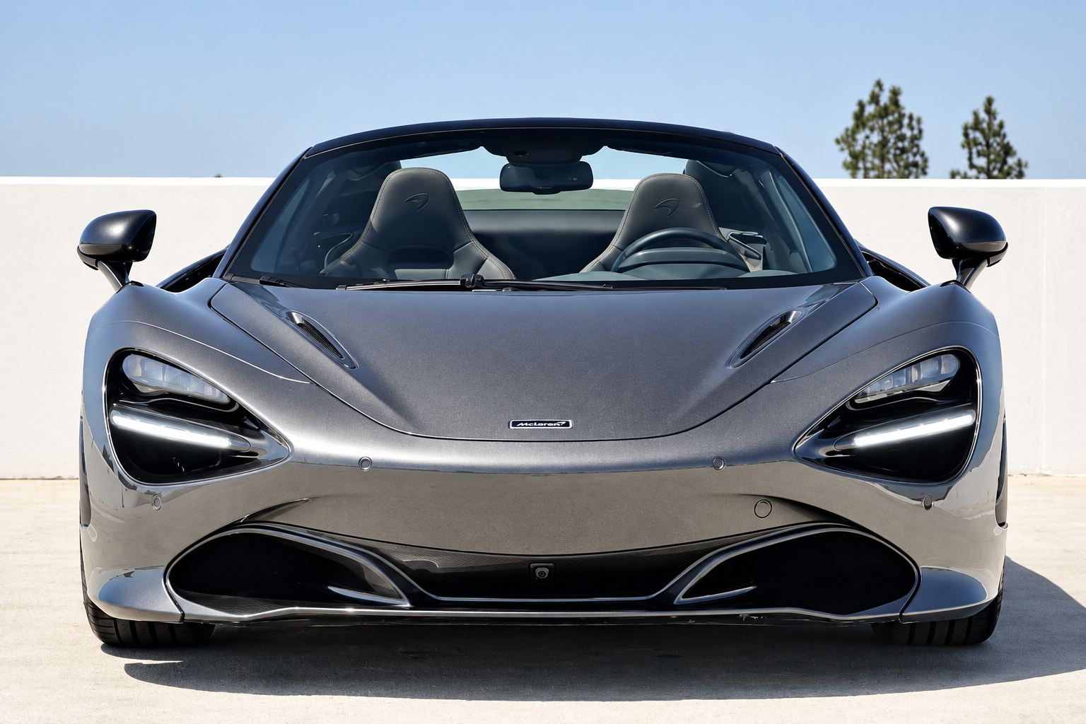 Head-on view of the dark grey McLaren 720S Spider, roof down, eye-socket headlamps and palm trees behind