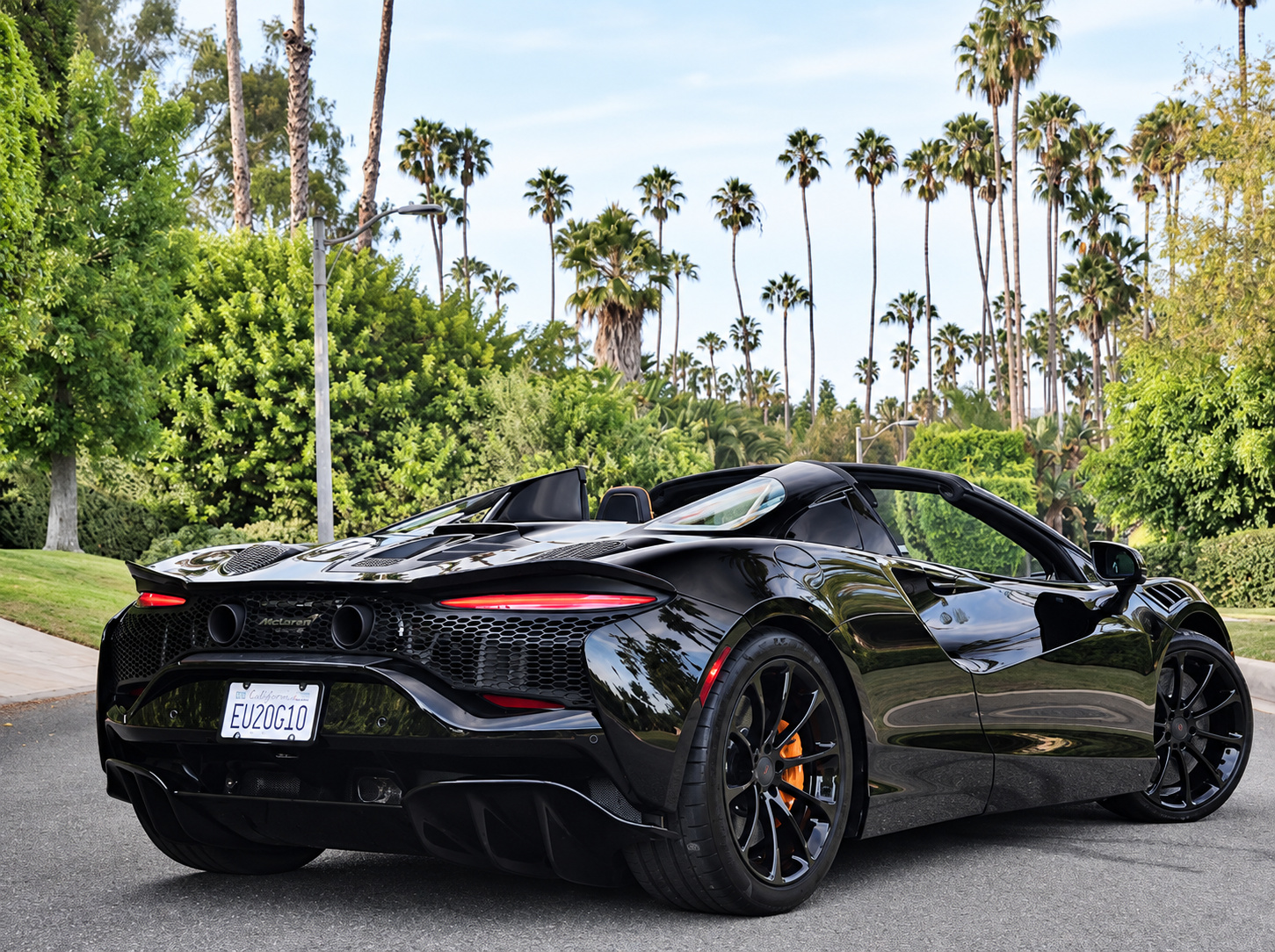 Rear three-quarter of the black Artura Spider, roof down, black wheels and orange calipers under a row of Beverly Hills palms