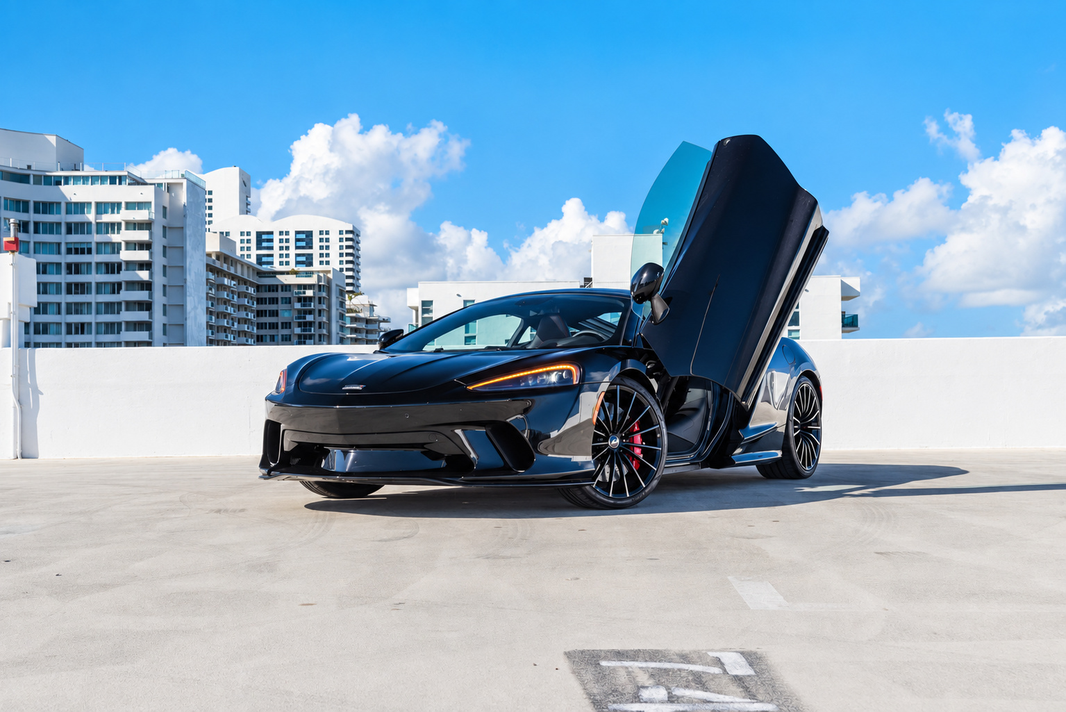 Black McLaren GT with the driver's door raised on a rooftop deck, white towers and blue sky behind