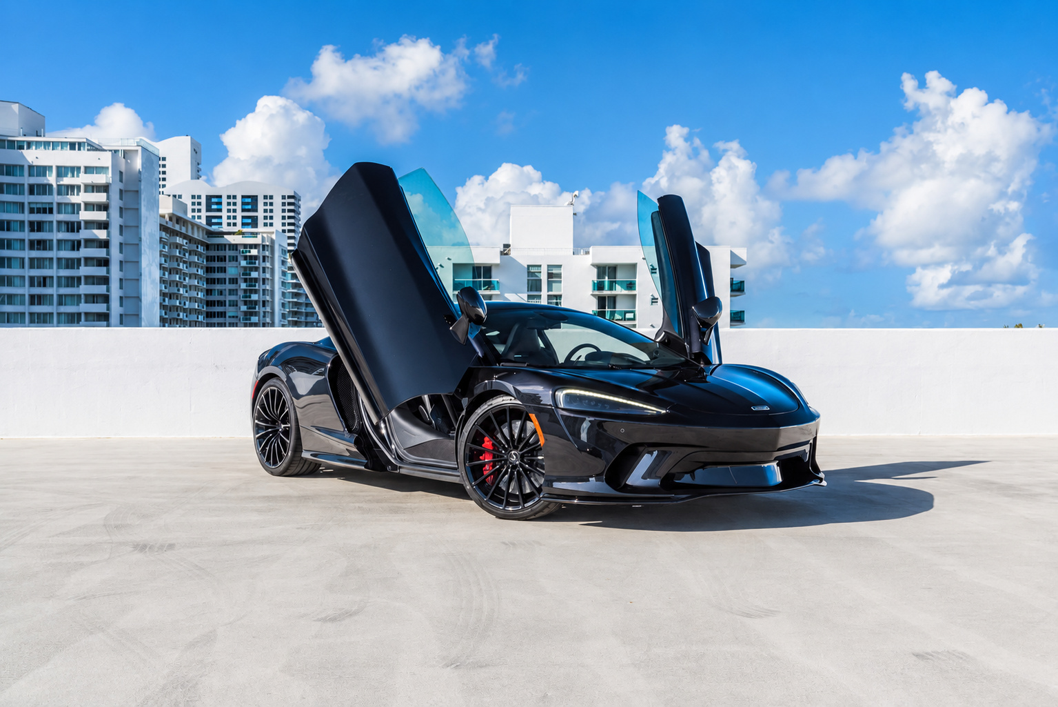 Black McLaren GT with both dihedral doors up, red brake calipers showing behind the black wheels