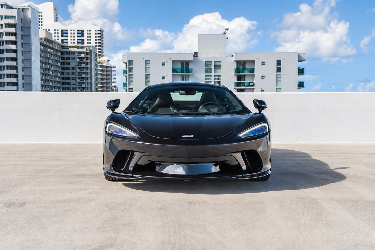 Head-on view of the black McLaren GT, low nose and headlamps, parked facing the camera on a rooftop deck