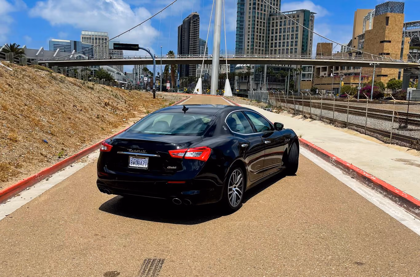 Rear three-quarter of the black Maserati Ghibli with a city skyline and a cable-stayed pedestrian bridge behind