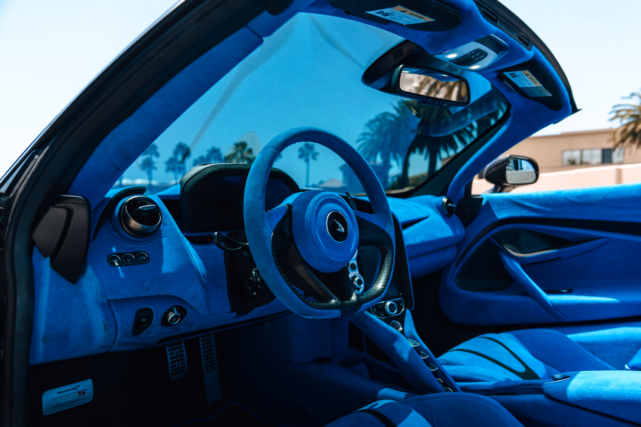 Driver's view of the 720S Spider, blue alcantara steering wheel and dash with the roof down and palms overhead