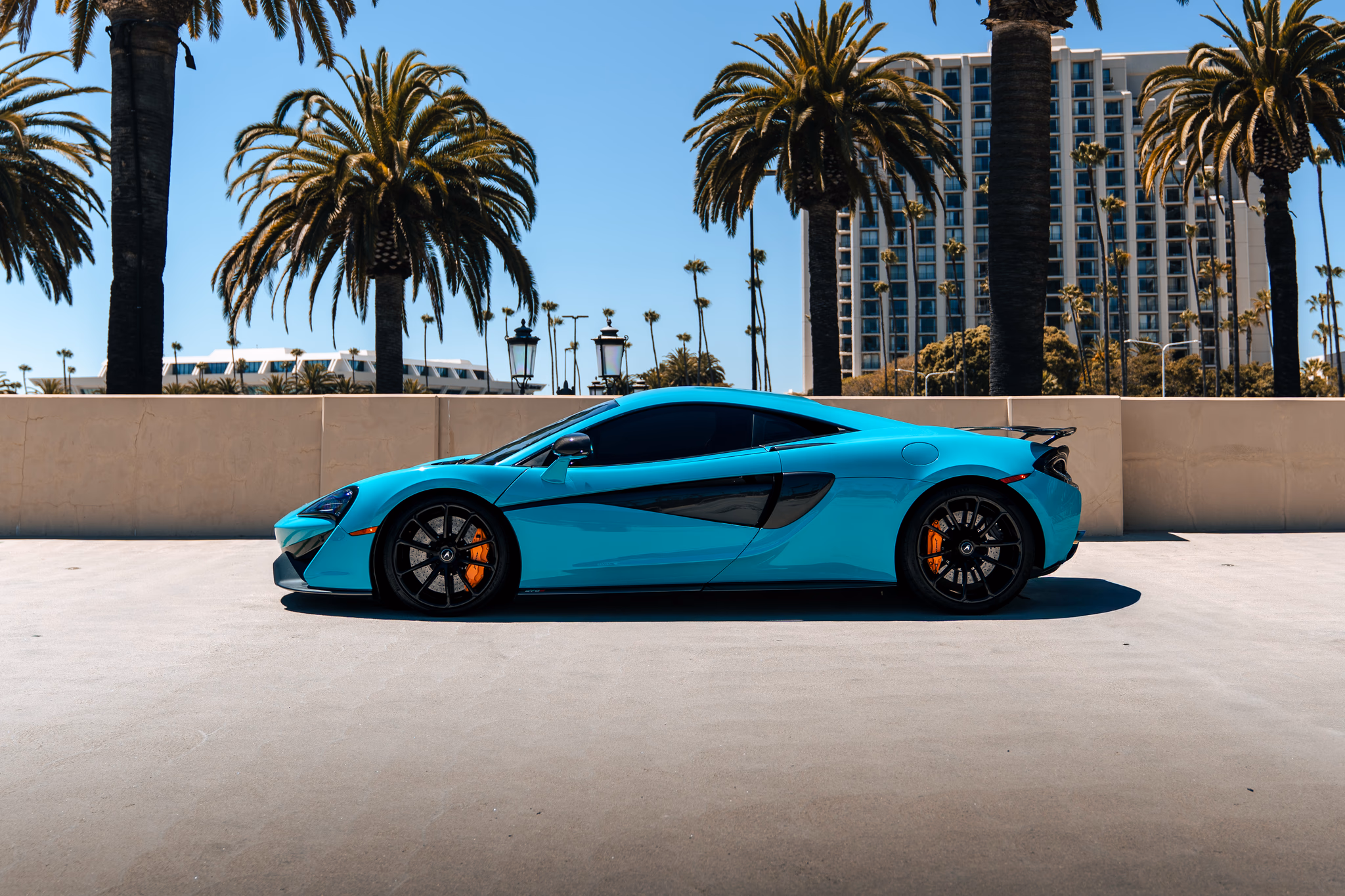 Side profile of the Curacao Blue McLaren 570S on black wheels with orange brake calipers, palms and a tower behind