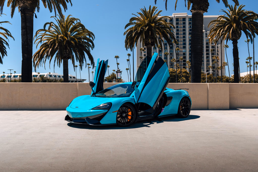 Curacao Blue McLaren 570S coupe with both dihedral doors raised on a palm-lined rooftop deck, towers behind
