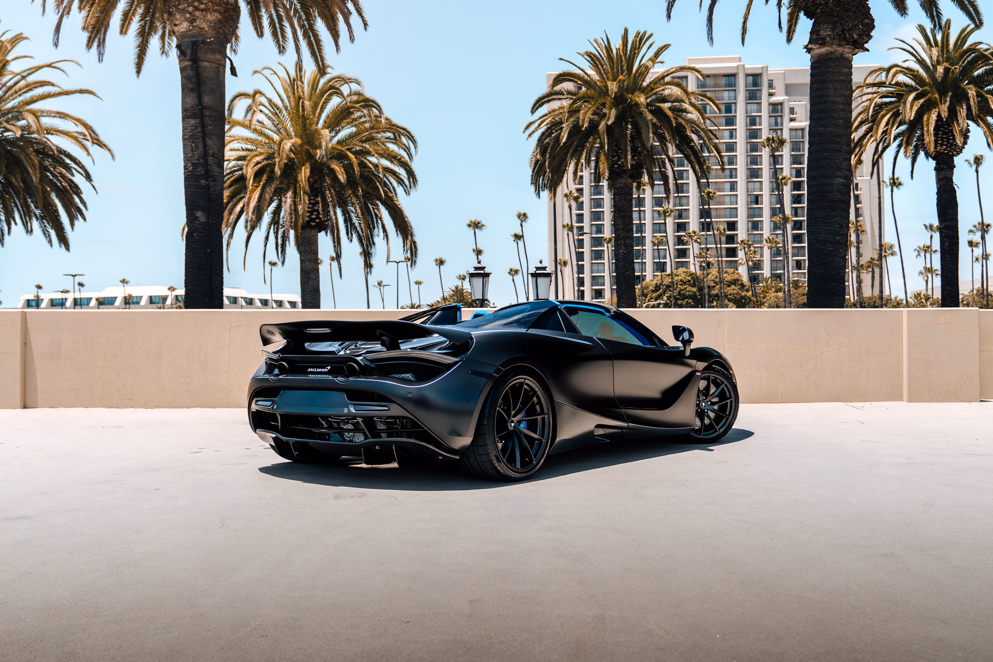 Rear three-quarter of the satin black McLaren 720S Spider, roof down, palm trees and a tower behind