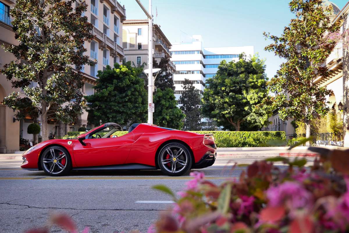 Side profile of the red Ferrari 296 GTS, roof stowed, on a Beverly Hills street with palms and a hedge behind