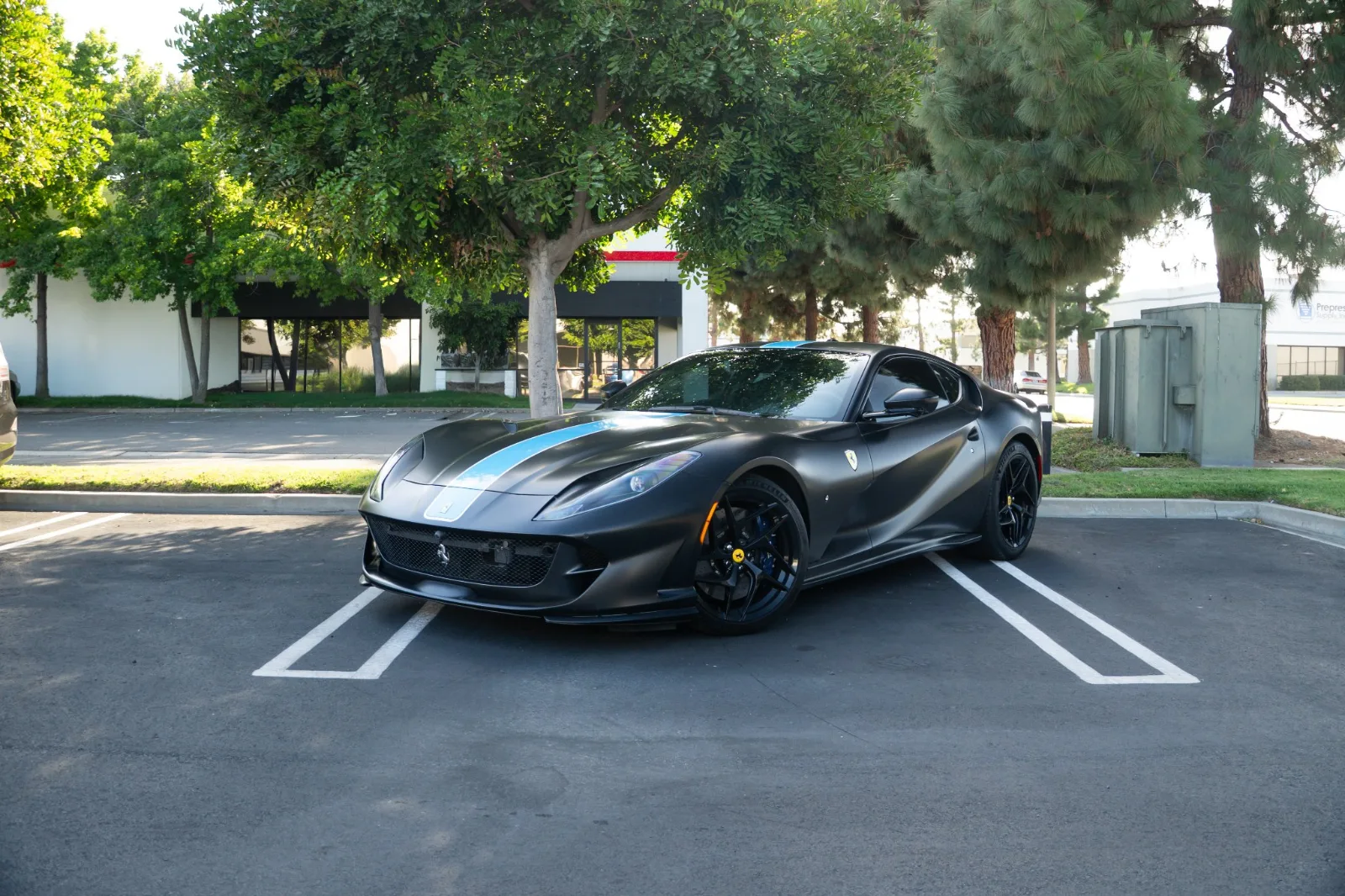 Satin black Ferrari 812 Superfast with a light blue centre stripe parked in an office lot under trees, front three-quarter