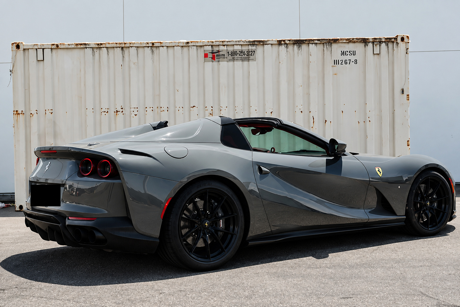 Rear three-quarter of the grey 812 GTS on black wheels, roof down, parked beside a shipping container