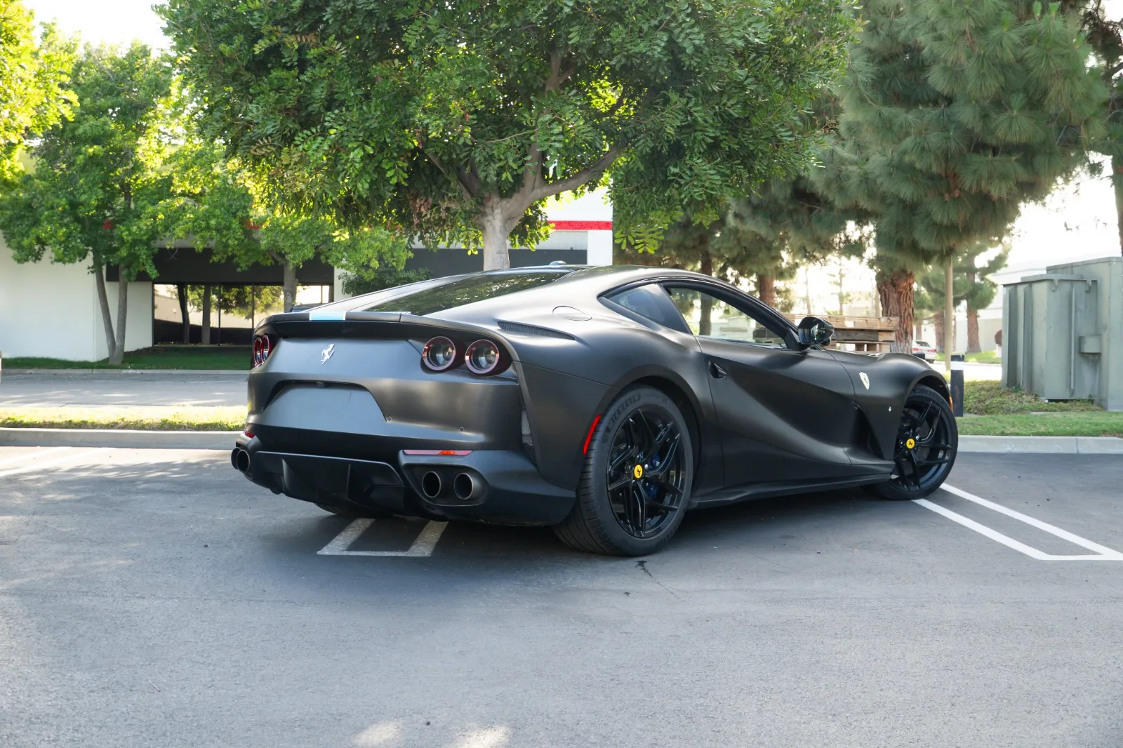 Rear three-quarter of the satin black 812 Superfast with the blue stripe, parked under trees in an office lot