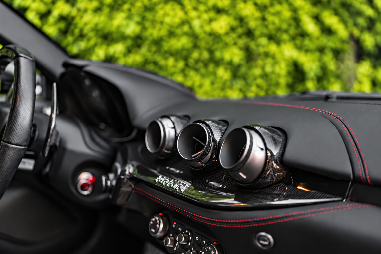 Centre of the Ferrari F12berlinetta dash with the 3 round vents and carbon trim, hedge visible through the glass