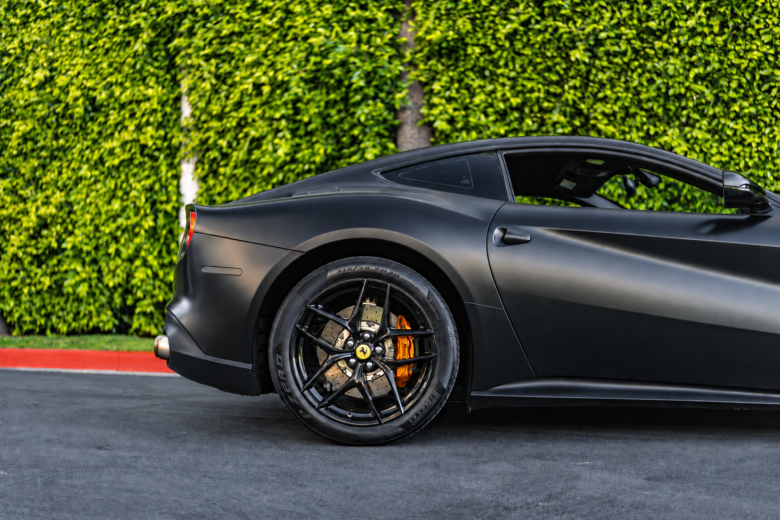 Rear quarter of the satin black Ferrari F12berlinetta against a green hedge, black wheel and yellow caliper