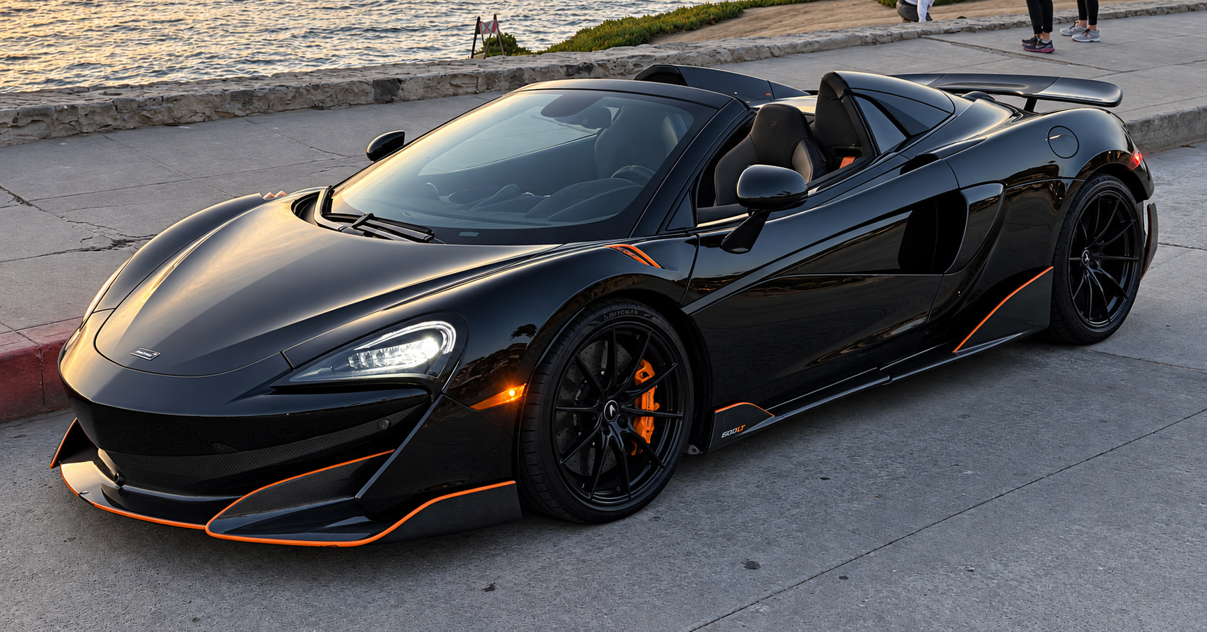 Front three-quarter of the black McLaren 600LT Spider with orange calipers and splitter accents, roof folded, hills behind