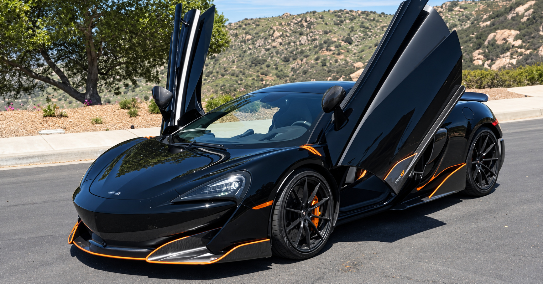 Black McLaren 600LT Spider with its dihedral door raised, orange pinstripe along the door blade, rock wall behind