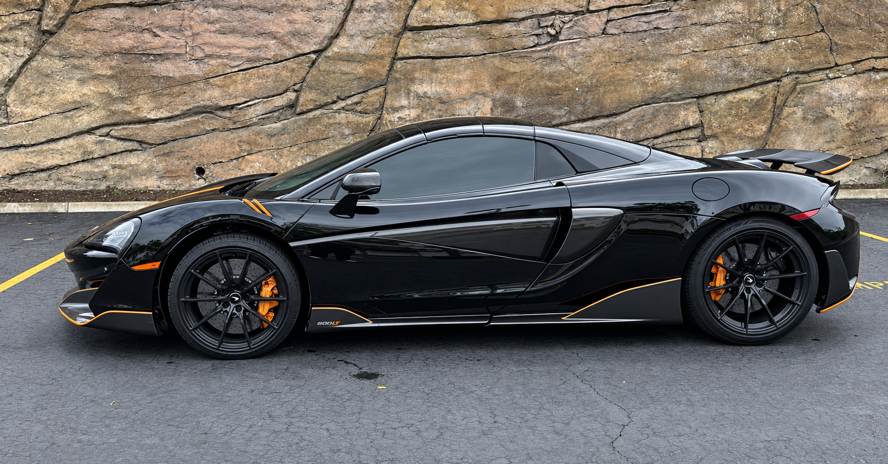 Side profile of the black McLaren 600LT Spider on black wheels with orange calipers, mountain rock wall behind