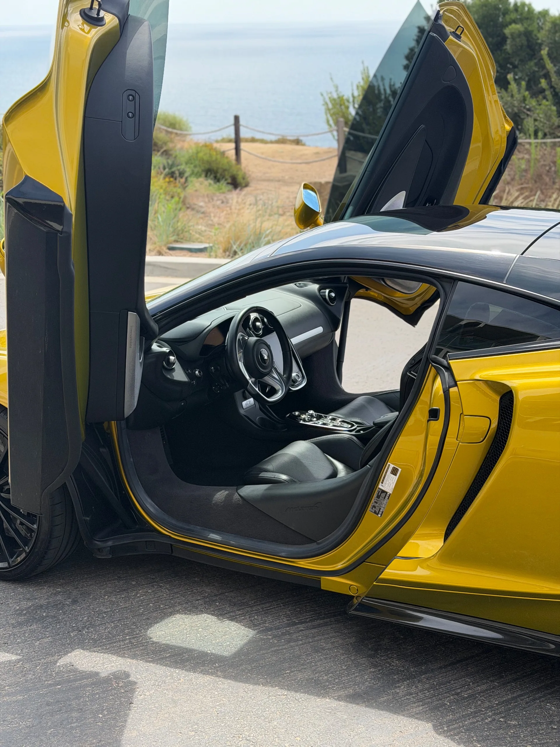 Open driver's door of the yellow McLaren GT showing the black leather cabin and steering wheel, La Jolla coast behind