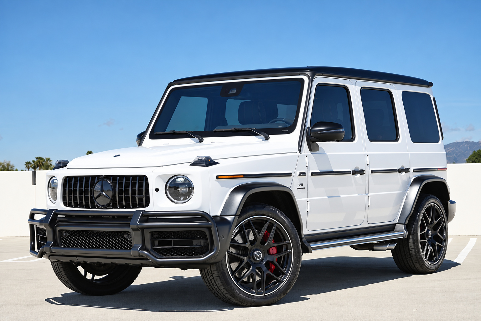 White Mercedes-AMG G63 with black wheels and red calipers, front three-quarter view on a rooftop deck with mountains behind