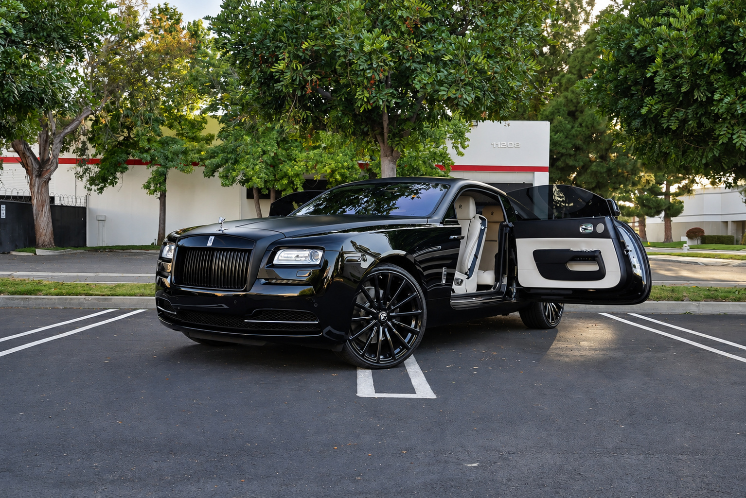 Black Rolls Royce Wraith with the coach door open showing the ivory cabin, parked in a tree-lined office park lot