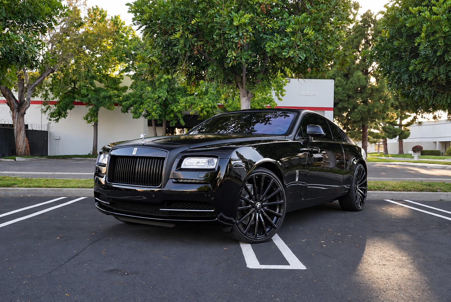 Front three-quarter of the black Rolls Royce Wraith, blacked-out grille and gloss black multi-spoke wheels, trees behind
