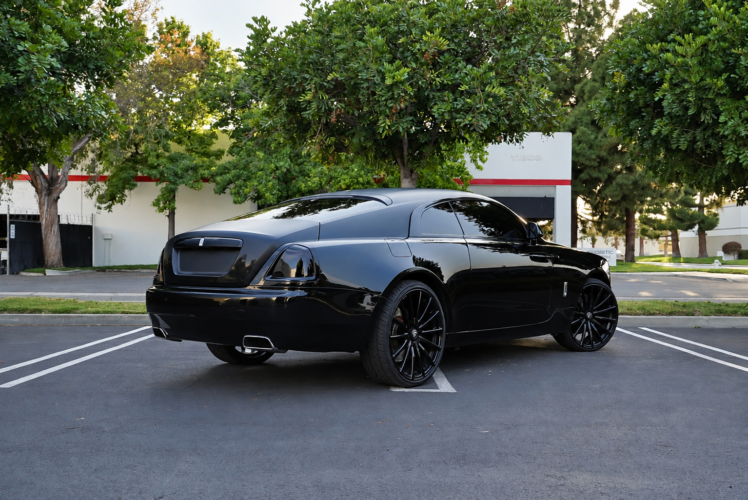 Rear three-quarter of the black Rolls Royce Wraith fastback under trees in an office park