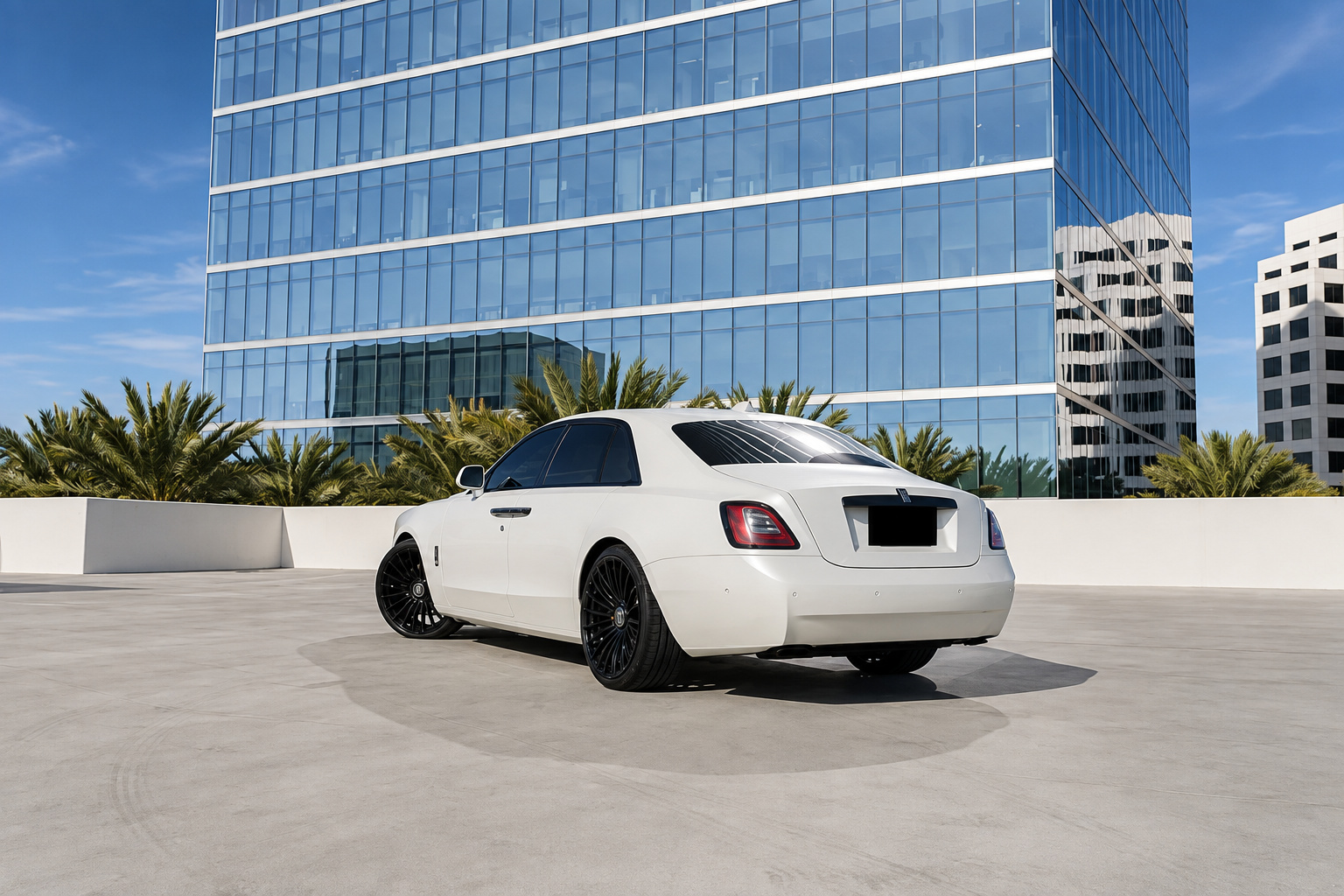 Rear three-quarter of the white Rolls Royce Ghost on black wheels with glass office towers behind