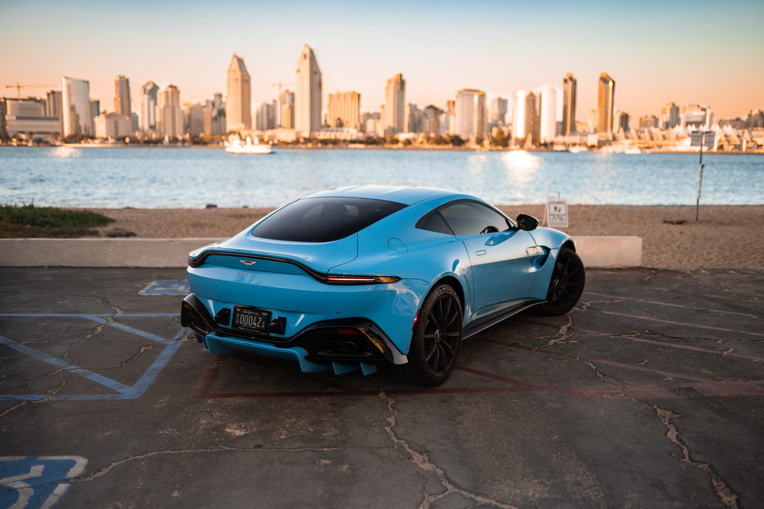 Rear three-quarter of the light blue Aston Martin Vantage on black wheels at golden hour, water and towers behind