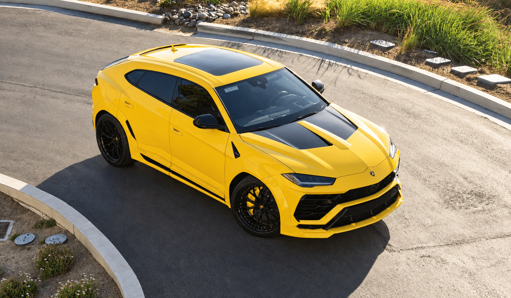 High angle of the yellow Lamborghini Urus Widebody on a curved road showing the black roof and hood accent