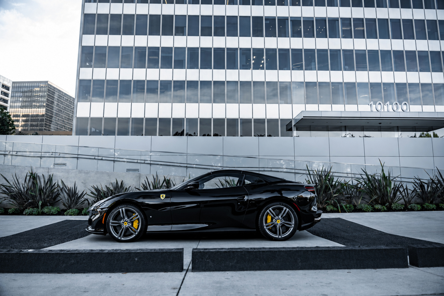 Side profile of the black Ferrari Portofino, roof stowed, silver wheels and yellow calipers, glass tower behind