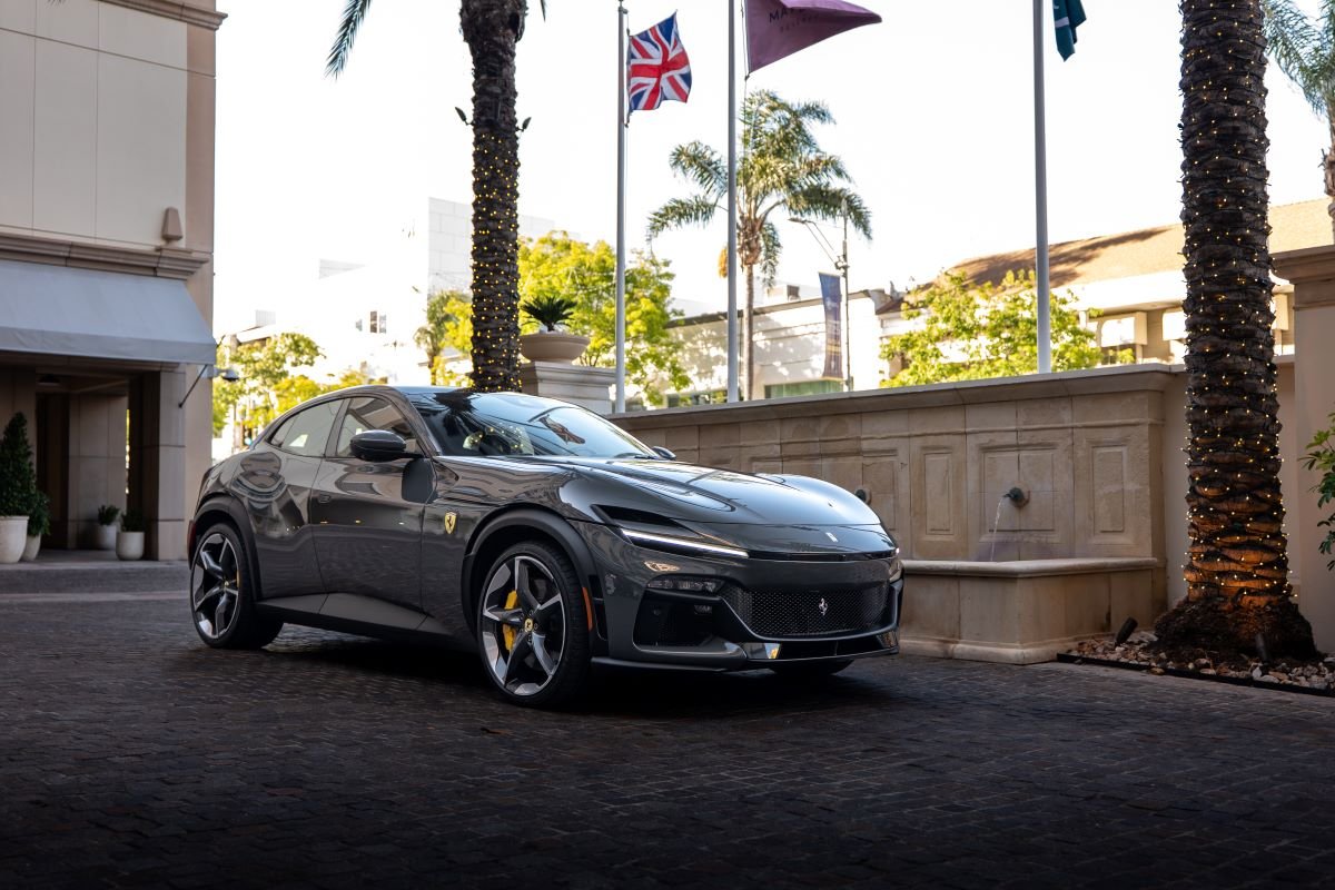 Dark grey Ferrari Purosangue parked at a hotel entrance under palms and flags, front three-quarter view