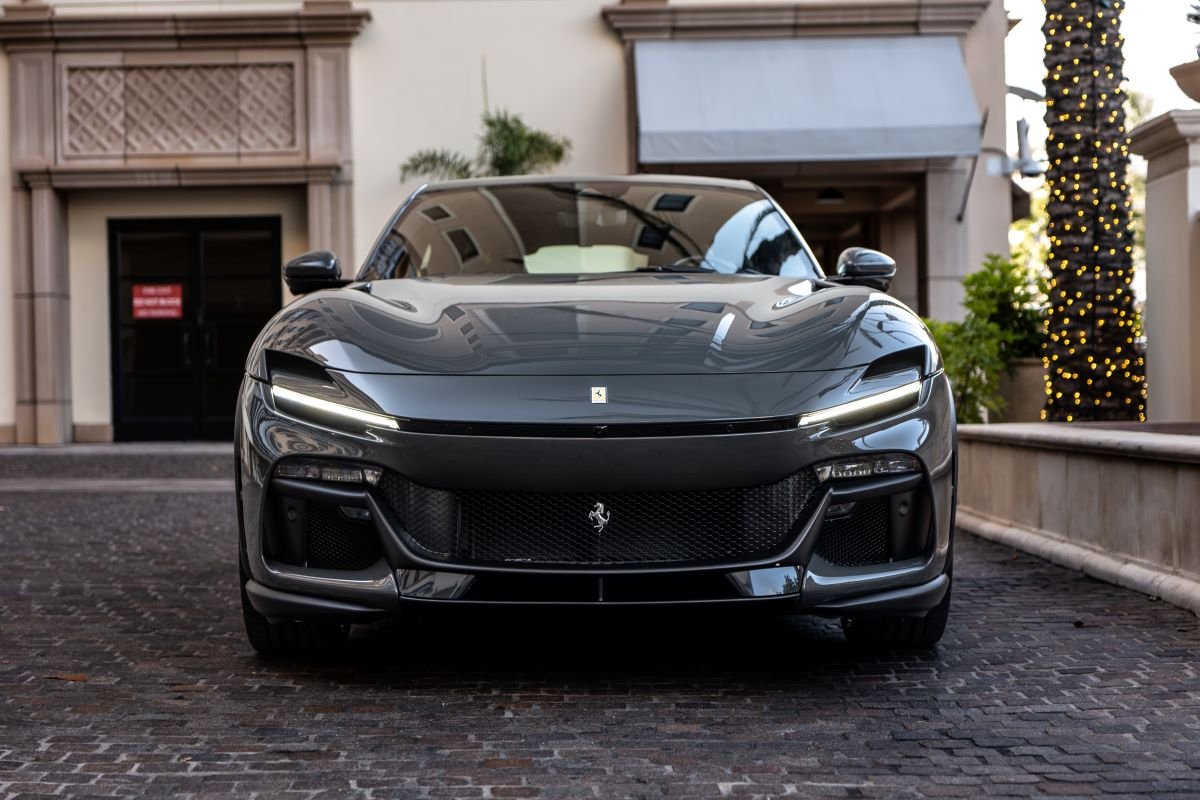 Head-on view of the dark grey Ferrari Purosangue on a cobbled hotel drive, lights on in the trees