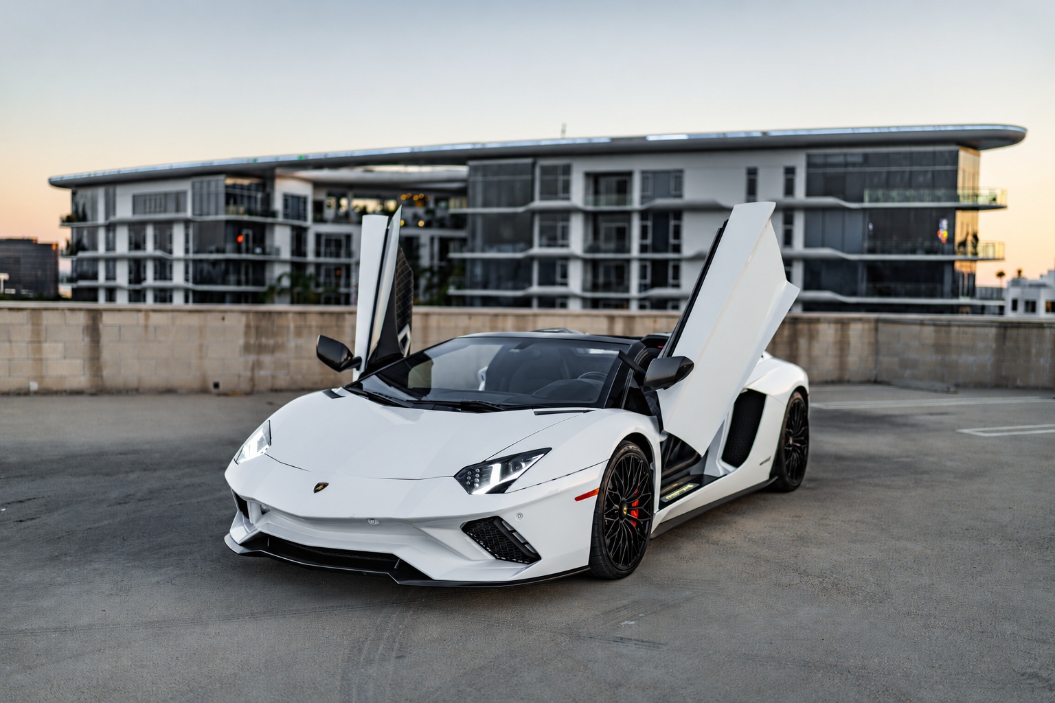 White Lamborghini Aventador S with both scissor doors up on a rooftop deck at dusk, modern apartment block behind