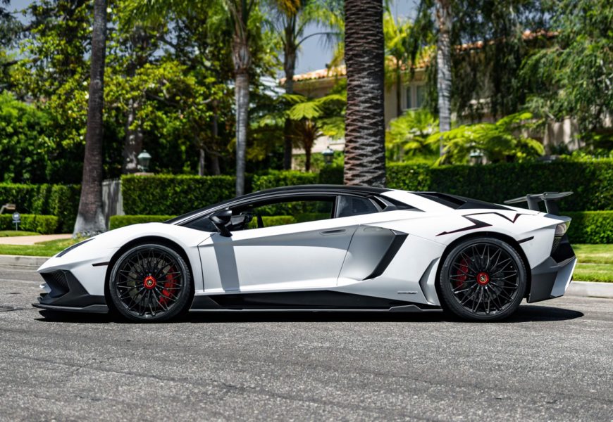 Side profile of the white Aventador SV with the black roof and carbon wing, black wheels with red calipers, hedges behind