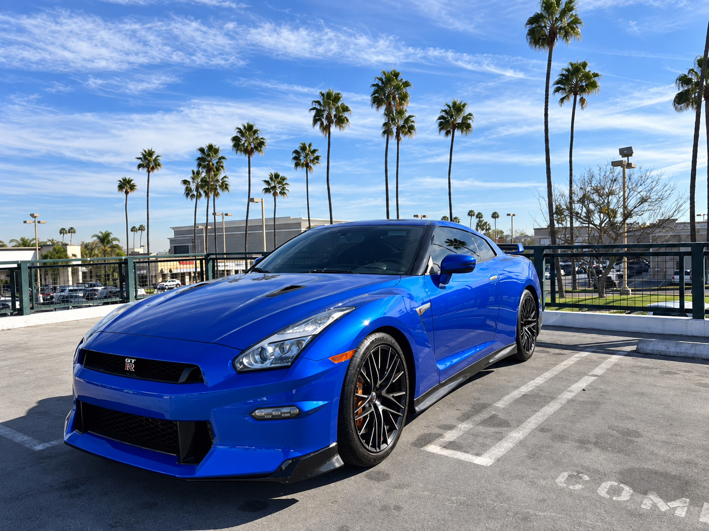 Front three-quarter of the Bayside Blue Nissan GT-R in a palm-lined lot, mesh grille and hood vents in the sun
