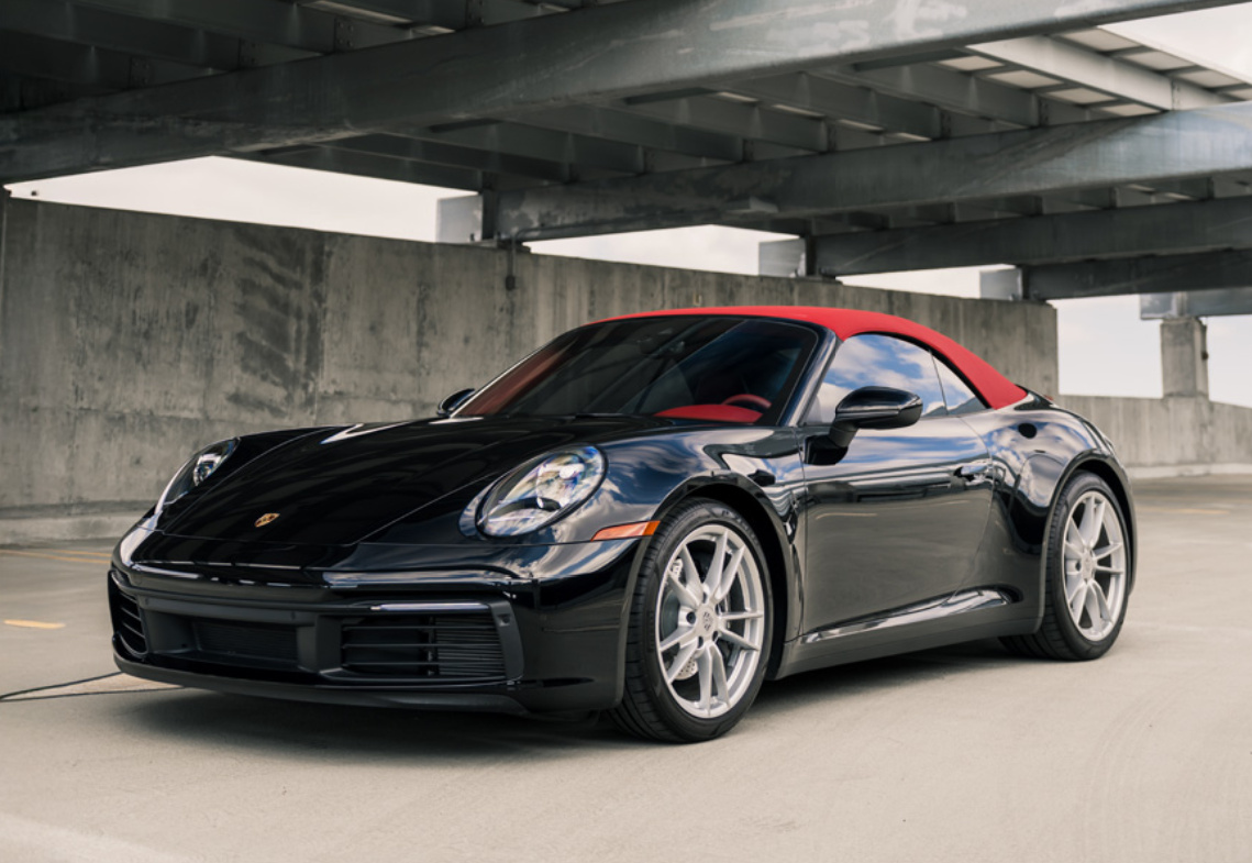 Black Porsche 911 Carrera Cabriolet with its red soft top up, front three-quarter view in a concrete parking garage