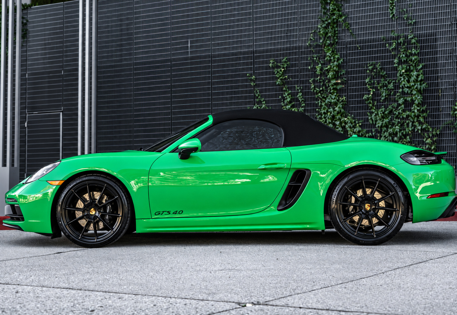 Side profile of the green Porsche 718 Boxster GTS 4.0, black top folded and black wheels, against an ivy wall