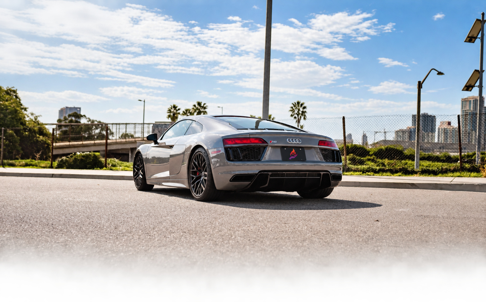 Rear three-quarter of the Nardo grey R8 V10 Coupe from a distance with palms and the San Diego skyline beyond the fence