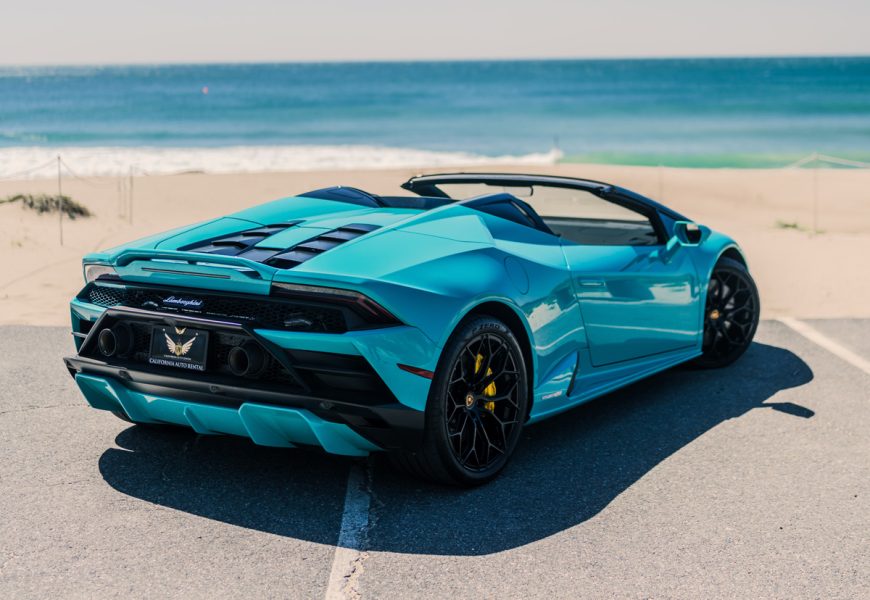 Rear three-quarter of the turquoise Huracan Evo Spyder, roof down, black wheels and yellow calipers with the Pacific behind