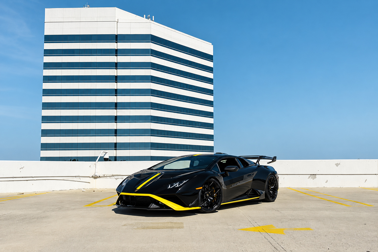 Black Lamborghini Huracan STO with yellow livery on an empty rooftop parking deck beside a striped office tower