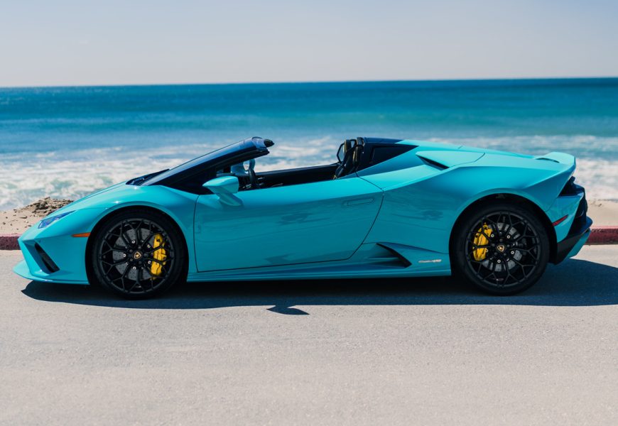 Side profile of the turquoise Huracan Evo Spyder, roof folded, black wheels, waves breaking behind on Zuma Beach