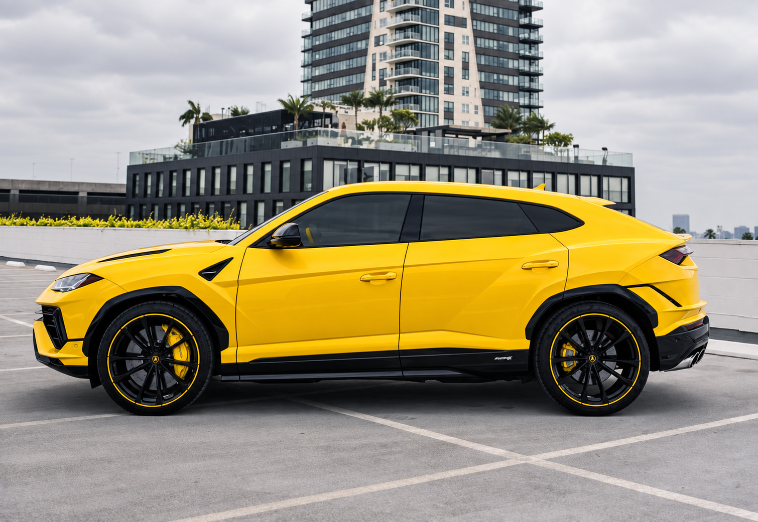 Side profile of the yellow Lamborghini Urus with black wheels and yellow calipers beside a glass tower