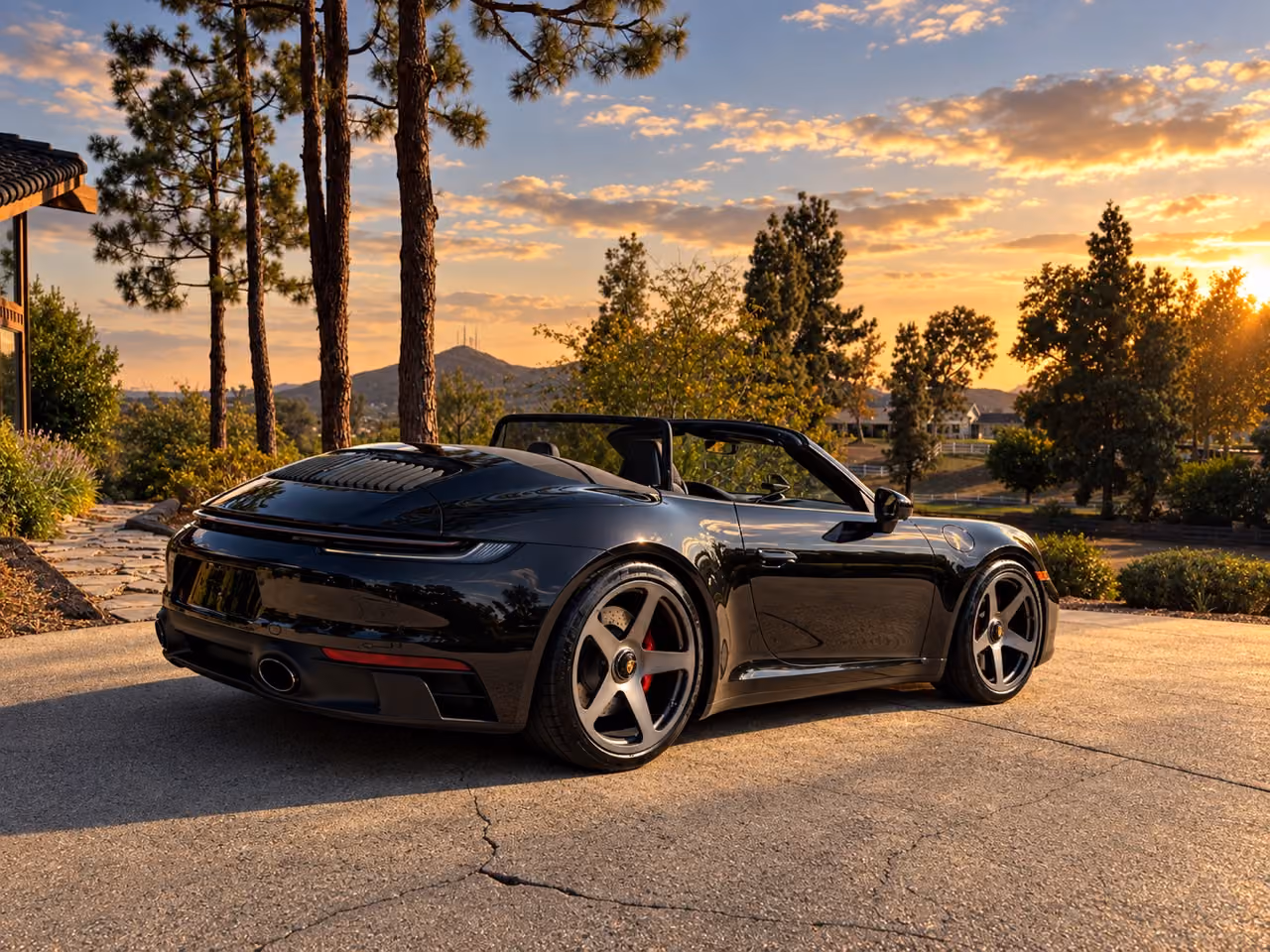 Black Porsche 911 Carrera GTS Cabriolet, top down, rear three-quarter in a driveway at sunset with pines behind
