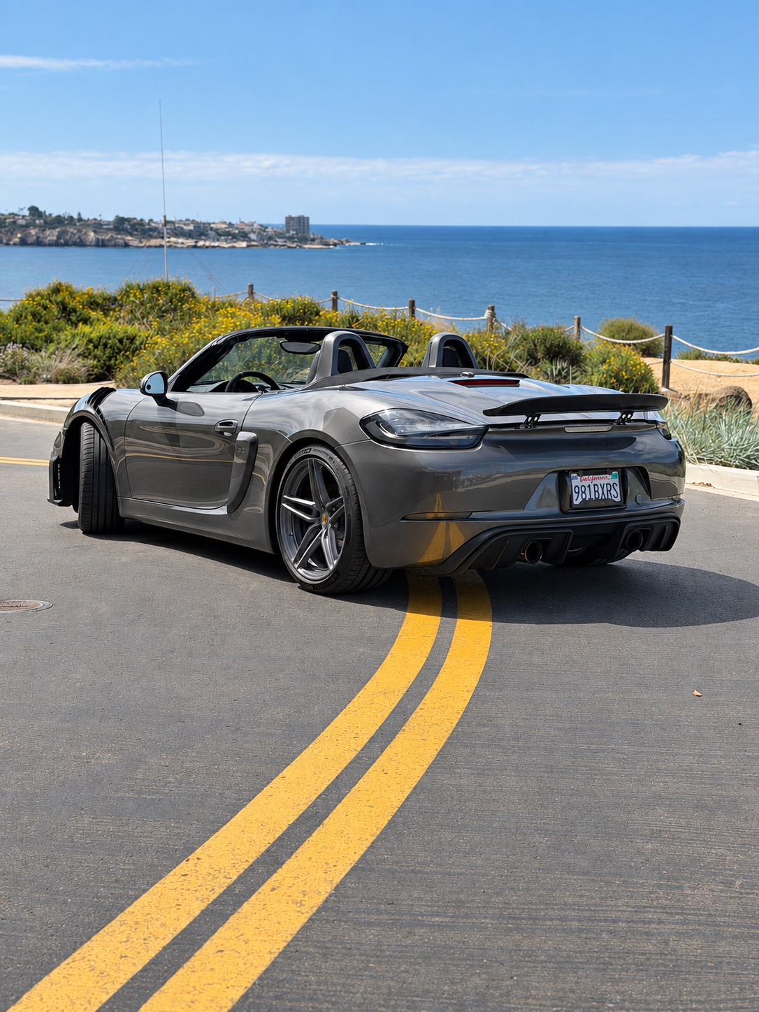 Rear three-quarter of the agate grey Porsche Boxster S, top down, carbon ducktail wing, ocean and cliffs of La Jolla behind