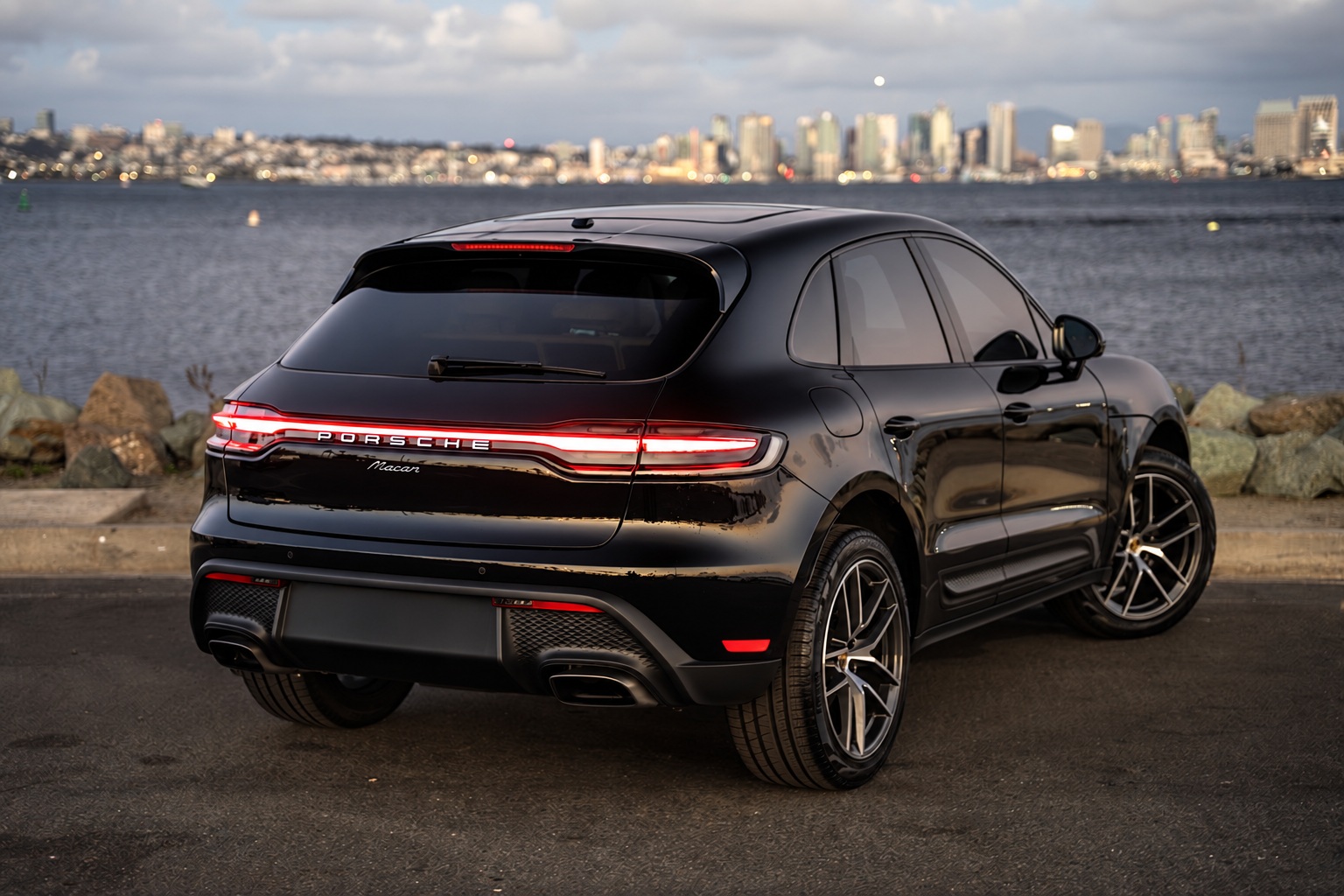 Rear three-quarter of the black Porsche Macan showing the full-width light bar, downtown San Diego across the bay